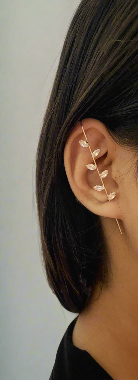 Close-up of an ear wearing a delicate gold earring with leaf-like designs against a neutral background.