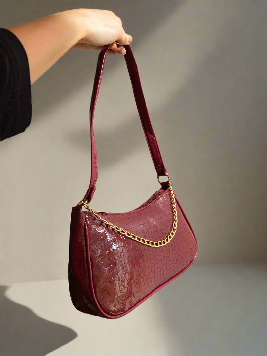 Maroon handbag with gold chain handle held by a hand against a plain background